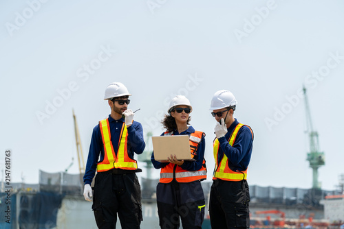 A group of engineers, wearing safety suits and helmets, are examining data on laptops in a shipyard and port area, with cranes and large ships in the background.