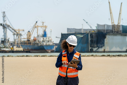 Portrait of female engineer in safety vest and helmet using radio and tablet at shipyard port, overseeing logistics and maritime shipping operations.