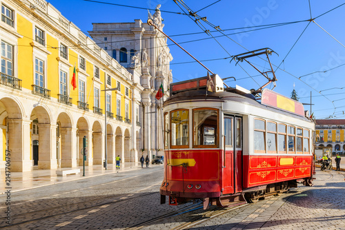 A classic red-and-white heritage tram rolls past the majestic Arco da Rua Augusta and grand yellow arcades of Praça do Comércio in the historic downtown core of Lisbon, Portugal.