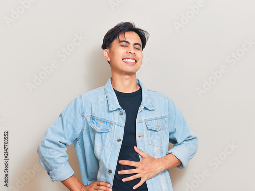 Happy young Asian man touching his stomach with eyes closed and smiling in satisfaction while standing against a studio background