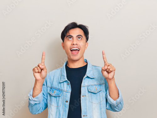 Excited young Asian man pointing both index fingers upward while looking up against a neutral studio background for advertising and promotional concepts.
