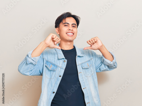 Obraz na plátně A proud young Asian man points both thumbs at himself while smiling at the camera in a studio setting wearing a denim jacket