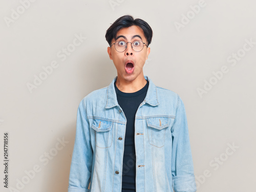фотографія Shocked young Asian man wearing eyeglasses and a denim jacket with a funny open mouth expression in a studio portrait against a neutral background