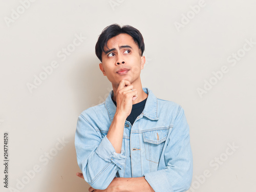 A pensive young Asian man touching his chin and looking up thoughtfully against a neutral studio background while considering an idea