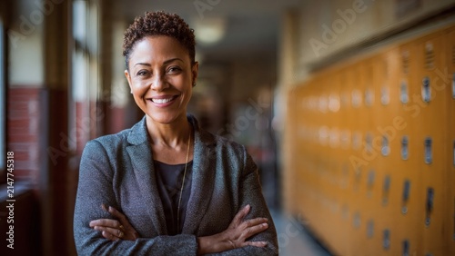 Female teacher smiling confidently while standing in a school hallway with lockers.