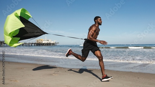 Foto Athletic Black man running on beach with resistance parachute training