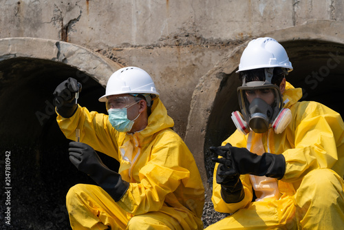 Environmental engineer in yellow hazmat suit and helmet collecting water samples from a drainage pipe for pollution and environmental quality inspection.