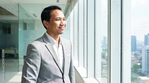 Indonesian business man investor, doubtful young male ceo executive wearing suit standing in office holding hand in chin looking away at work thinking on leadership question, feeling doubt.