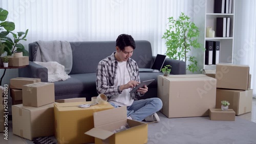 Young Asian man managing online business orders with tablet surrounded by shipping boxes in modern apartment