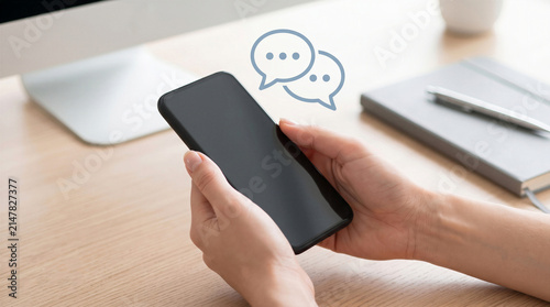 Person holding smartphone with blank screen and chat bubbles above it at office desk. Online messaging and communication concept