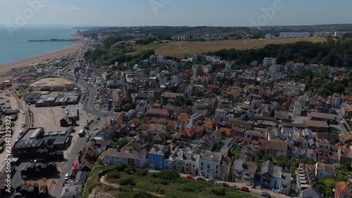 Left aerial orbit sweeps over Hastings rooftops, with the net sheds, East Hill, West Hill and Hasting Pier visible in the distance, with clear blue sea and blue summer skies.