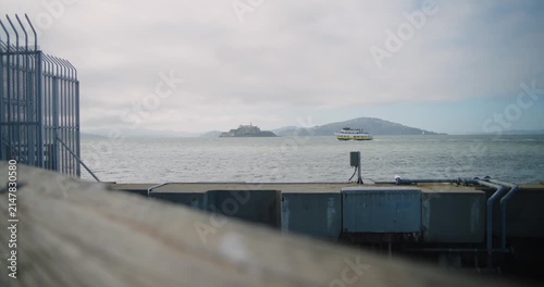 4K handheld cinematic view of boat passing by Alcatraz Island from the San Francisco waterfront near Pier 39. The historic prison island sits across the bay with surrounding water. Pier in foreground.