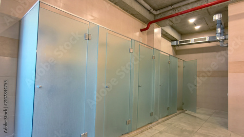 Frosted glass stalls line a public restroom, providing privacy within a clean and modern facility featuring concrete ceilings, exposed pipes, and ventilation systems
