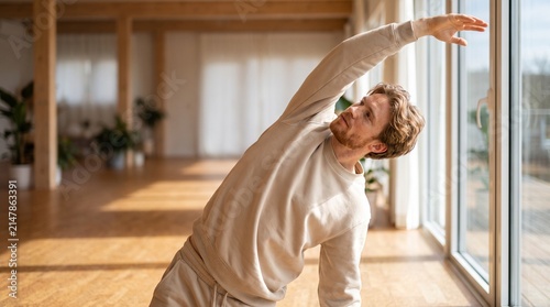Young man stretching side body during indoor yoga practice in bright wooden studio.