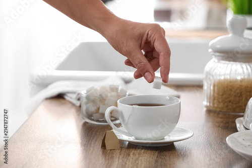 Fotografie Hand adding a white sugar cube to a cup of hot black tea on a wooden kitchen cou