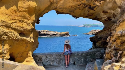 woman tourist wearing hat looking through natural rock arch at ocean and biarritz lighthouse in france