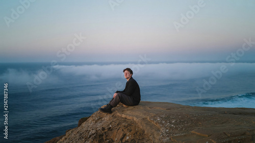 Man sits alone on the edge of a rugged coastal cliff overlooking the vast dark ocean during a misty twilight hour while contemplating nature in a moment of quiet solitude and personal reflection.