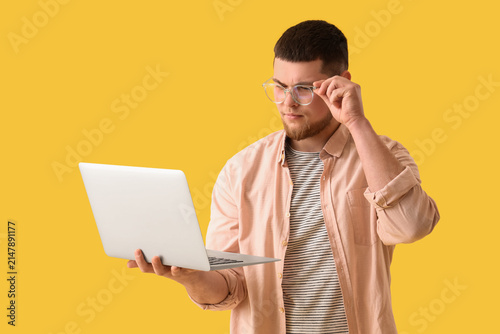 Young man in eyeglasses using laptop on yellow background