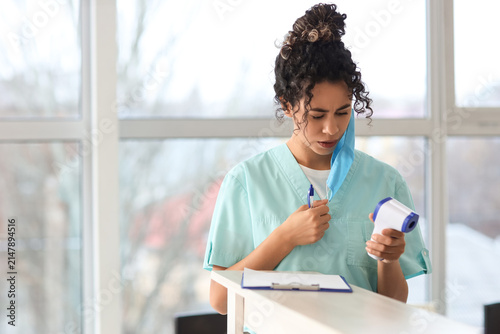 Female African-American medical intern with mask and infrared thermometer in ...