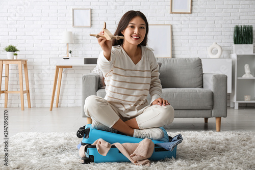 Young woman with wooden plane sitting on suitcase at home