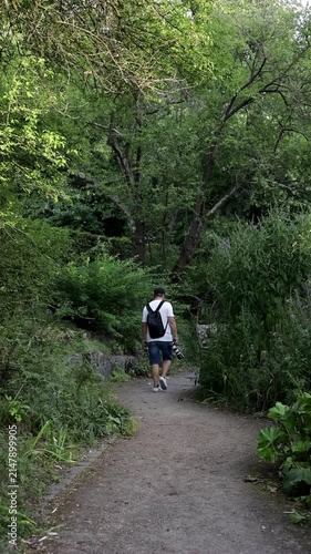 A man carrying a camera walks away along a lush garden path and disappears behind dense bushes, illustrating travel, exploration, photography, and outdoor leisure.