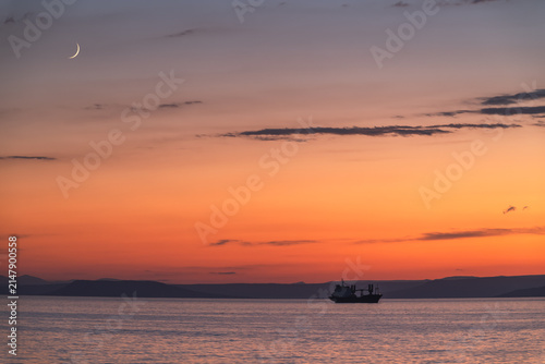 Silhouette of a cargo ship on a calm sea against a colorful sunset with a crescent moon.