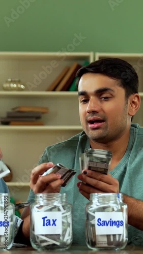 Indian Asian young couple in twenties sitting at home desk, placing Indian currency notes in three glass jars labeled Gold, Mutual Funds, Fixed Deposit, symbolizing savings, investment, financial plan