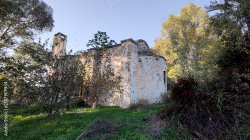 Canvastavla Exterior view of the historic Acendu Church (Agios Georgios) in Lefke, showing its stone apse and bell tower surrounded by lush green trees and foliage in warm sunlight