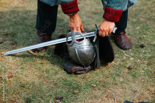 Foto A man in medieval clothing kneels on grass, handling a sword and helmet near a leather piece