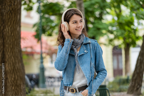 Young woman listening to music walking in city street smiles with headphones and enjoys relaxing moment in park. Girl tourist feels happy outdoors and looks up with joy while hearing favorite song.