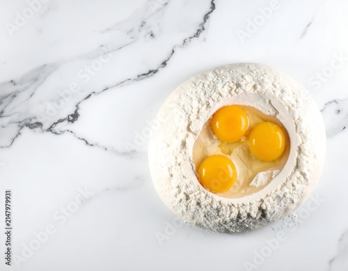 Top-down view of flour with three eggs in the center, on a marbled surface