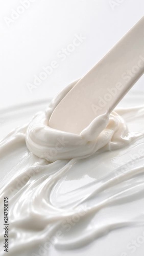 Smooth white cosmetic cream being mixed and scooped with a spatula on a glass surface. Macro close-up of luxury moisturizer lotion texture, dermatological beauty product, and hydrating skincare concep