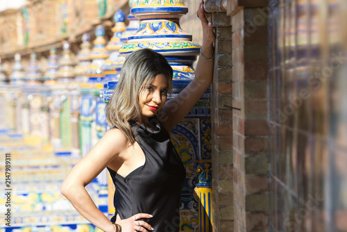 A young, beautiful woman leaning against the tiled wall of the famous Spanish Square in Seville. She is resting and smiling, enjoying the sunny day. Travel and holidays concept.