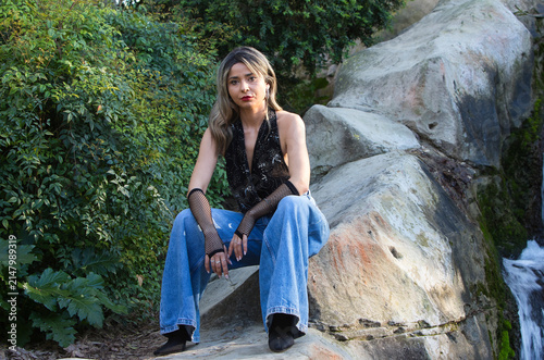 A young, pretty woman, wearing make-up and dressed in jeans, gloves and a black T-shirt, sitting on the rocks beside a crystal-clear waterfall surrounded by lush vegetation.