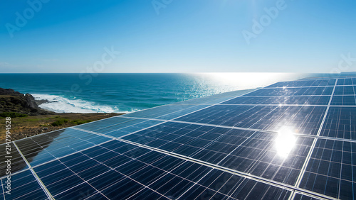 Solar panels array on a cliff overlooking the ocean with bright sun reflection, renewable energy concept