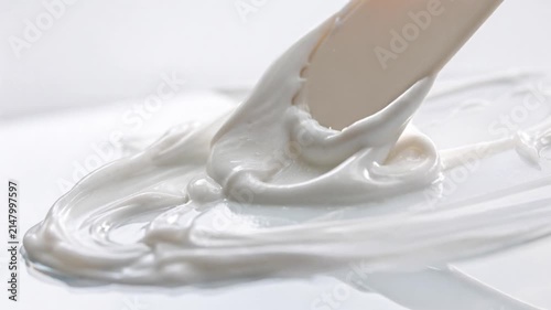 Smooth white cosmetic cream being mixed and scooped with a spatula on a glass surface. Macro close-up of luxury moisturizer lotion texture, dermatological beauty product, and hydrating skincare concep