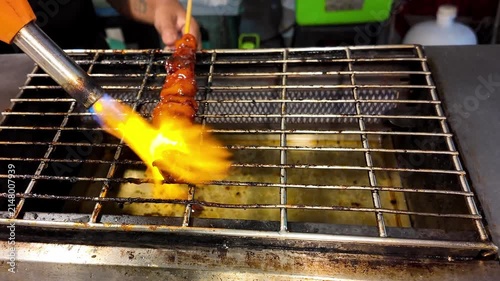 Close-up of a street food vendor using a blowtorch to sear a crocodile meat skewer on a grill at a Thai night market. Authentic exotic street food cooking with open flame, grilled crocodile meat and t