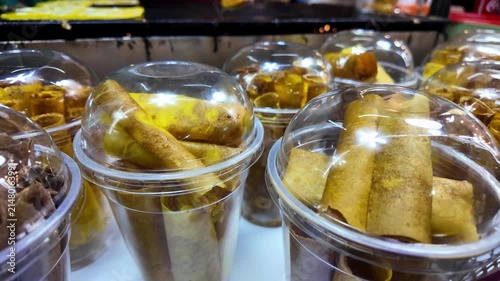 Packaged Thai crispy coconut wafer rolls displayed in clear takeaway cups with lids at a busy night market. Traditional coconut dessert freshly prepared and ready for customers to enjoy or take away. 