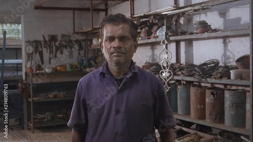 Middle aged Indian skilled, confident male fabricator worker in a uniform, standing and facing or looking at the camera in a fabrication workshop surrounded by the tools, equipment, and raw materials