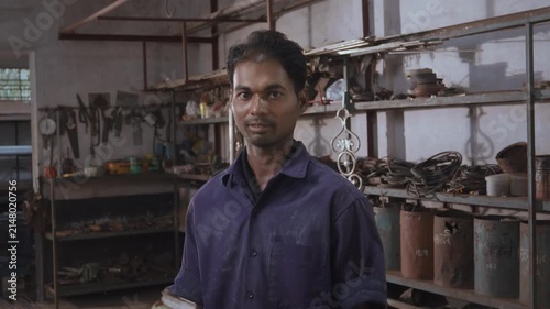 Young Indian skilled, confident, and happy male fabricator worker in a uniform, standing and facing or looking at camera in a fabrication workshop surrounded by the tools, equipment, and raw materials