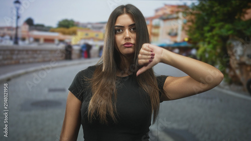 Konstfotografi Young hispanic woman showing thumbs down gesture on a cobblestone street with stone wall and lamppost in view; disapproval dismissal