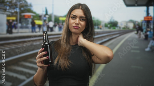 Obraz na płótnie Woman holding beer bottle in one hand and touching her neck with the other on a street train platform, gazing with a resentful expression; boredom
