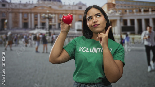 Konstfotografi Young volunteer woman in green shirt holds red heart toy aloft while touching chin in front of building; compassion reflection