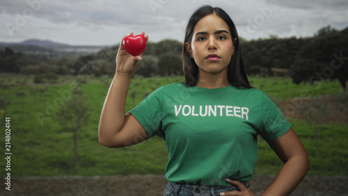 Fotografie Woman volunteer in green shirt holding red heart aloft with raised hand, hand on hip and eyes closed in a forest; compassion charity solidarity