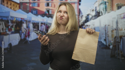 Foto Woman holding smartphone and paper bag on street, purses lips in a dismissive pout; frustration waiting