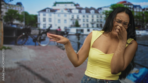 Foto Woman pinched nose with hand and squinted while holding face beside canal bicycles on a city street; disgust odor discomfort