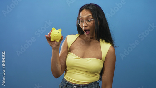 Photography Woman holding a yellow piggybank in her right hand with mouth open in surprise, arm extended and glasses visible, studio setting; saving optimism