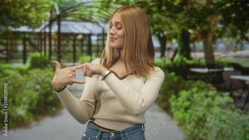 Fotografie Woman redhead showing call me hand gesture with hand to ear on a tree lined street cafe path; playful invitation