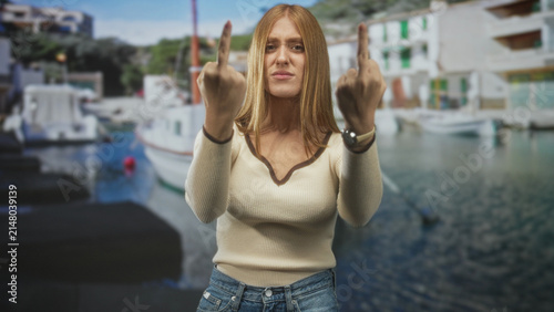 Quadro su tela Woman holding up middle fingers at a harbor pier, redhead in cream sweater and denim jeans with wristwatch visible; defiance attitude