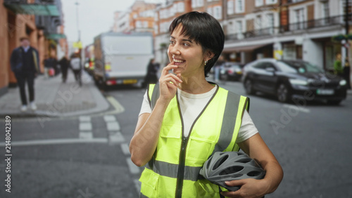 Konstfotografi Woman with short hair in high vis vest smiling while holding bicycle helmet with hand on chin on street; thoughtful reflection